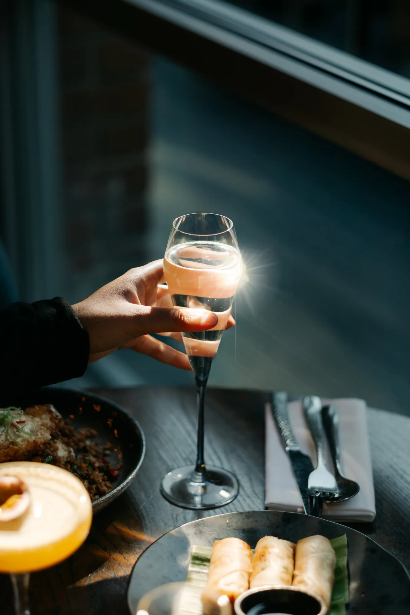 A glass of Prosecco being raised over a Pornstar Martini and spring rolls